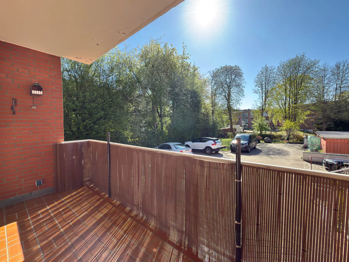 Balcony view with brick wall, bamboo fence, and tiled floor. Cars and trees are visible in the background under a sunny sky.