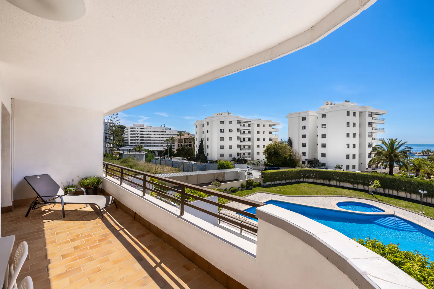 Balcony view of a pool and white buildings. A lounge chair sits on the brick patio under a white ceiling. Blue sky.