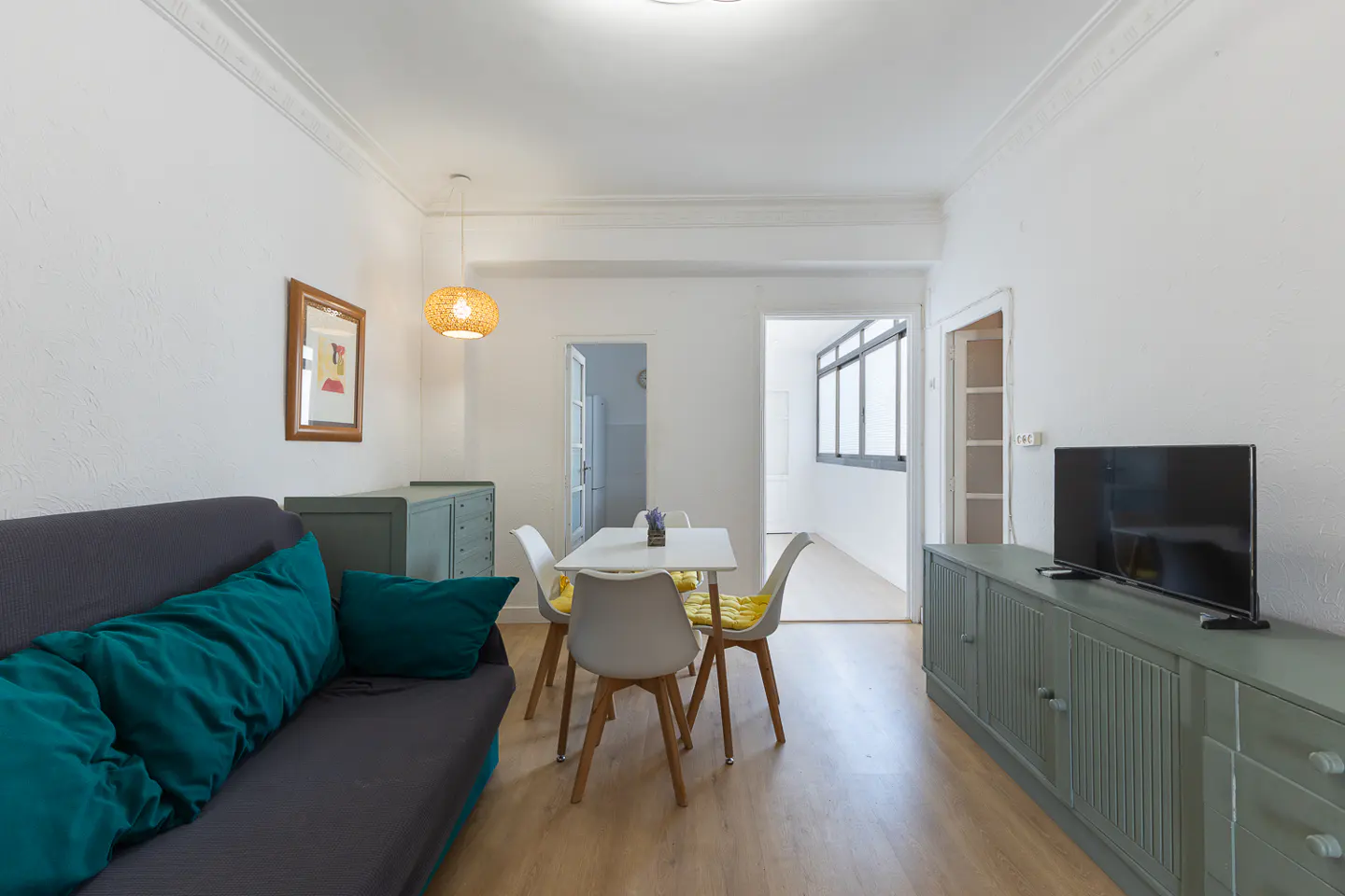 Bright living room with gray sofa, teal pillows, white walls, wood floors, and a dining table with four chairs. A TV sits on a green cabinet.