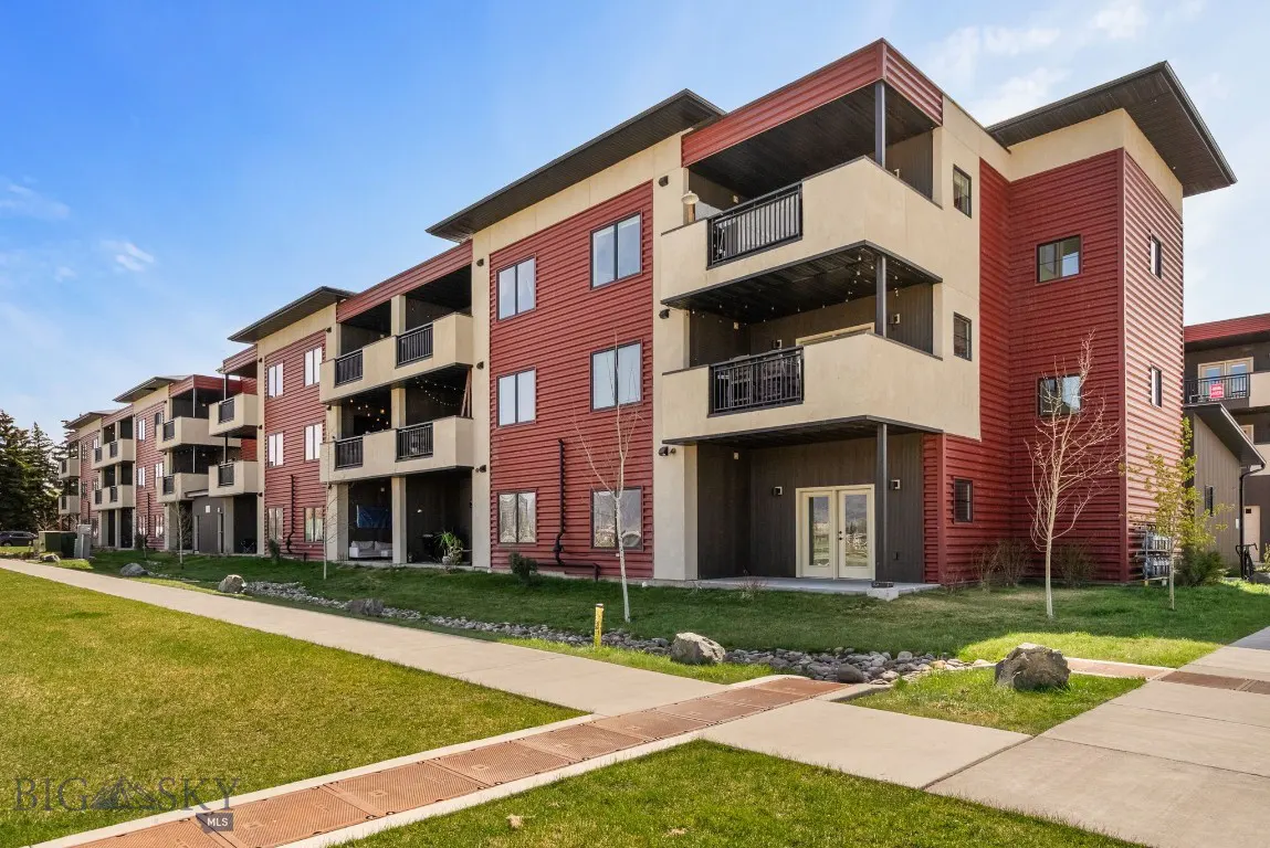 Modern three-story apartment building with red siding, beige accents, and black balconies under a blue sky.