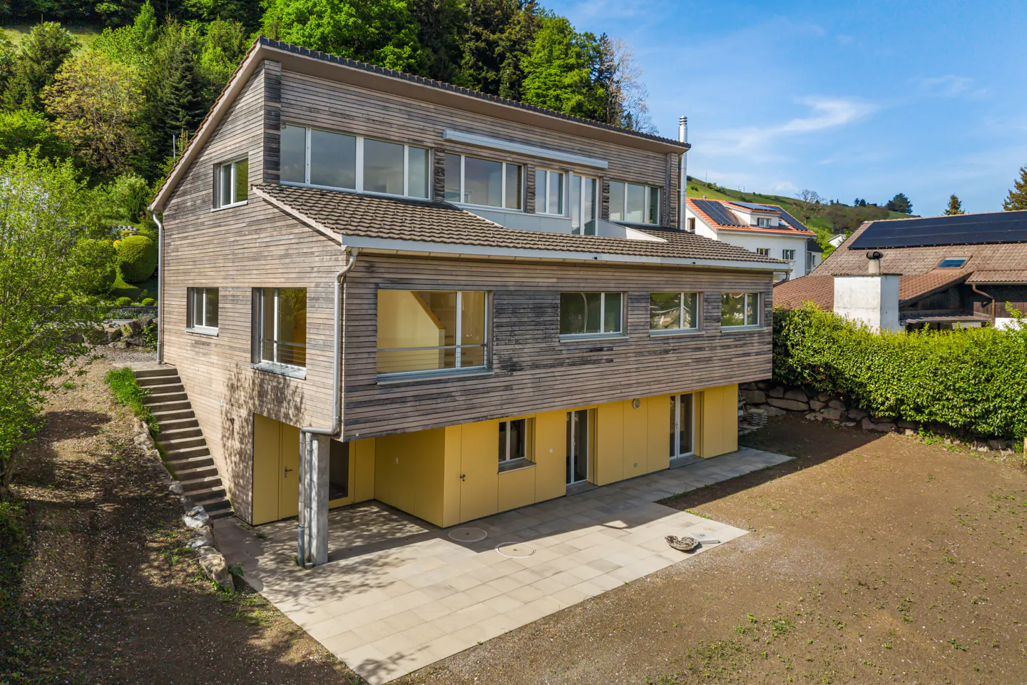 A modern, three-story house with wood siding and a yellow foundation, set against a green hillside. A stone patio extends from the house.