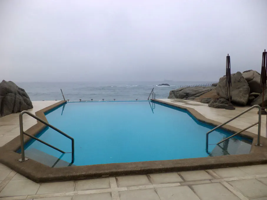 Outdoor pool with blue water and metal railings, overlooking a gray ocean and rocky coastline under an overcast sky.