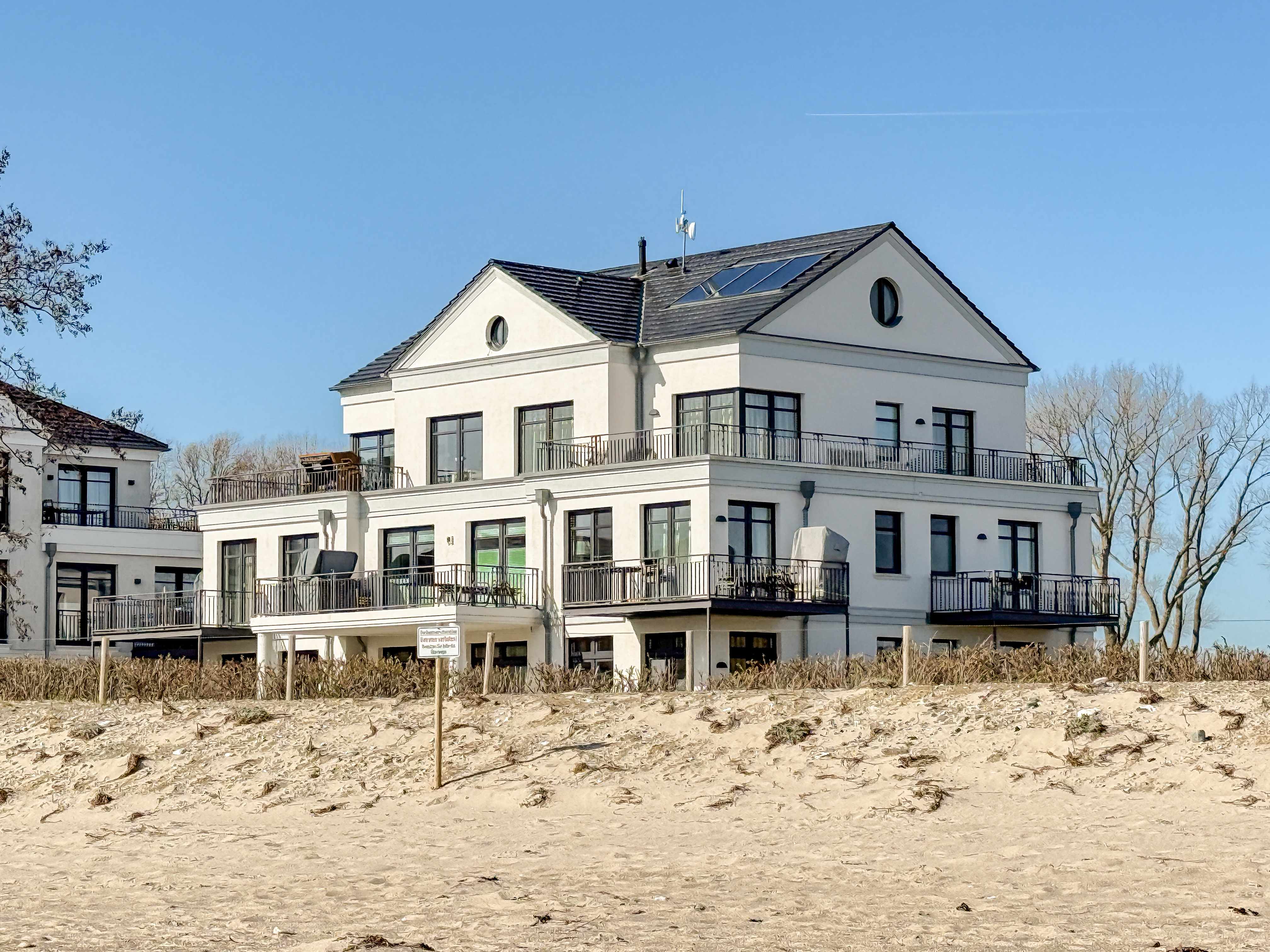 Three-story white building with black trim and balconies, set on a sandy beach under a clear blue sky.