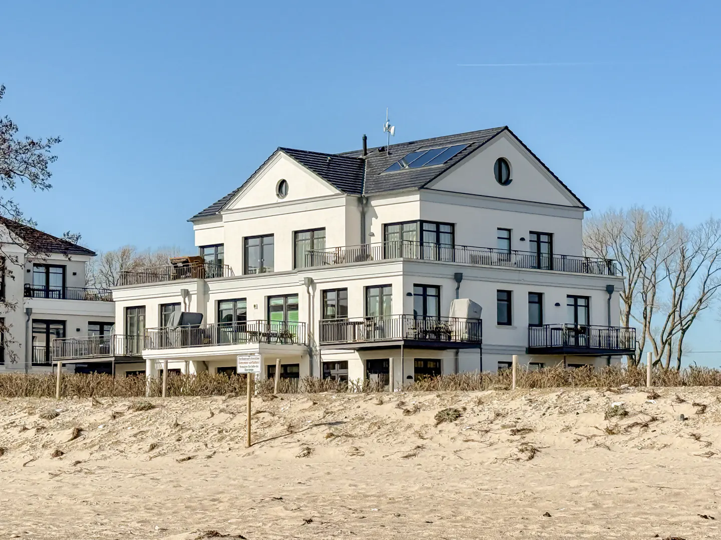 Three-story white building with black trim and balconies, set on a sandy beach under a clear blue sky.