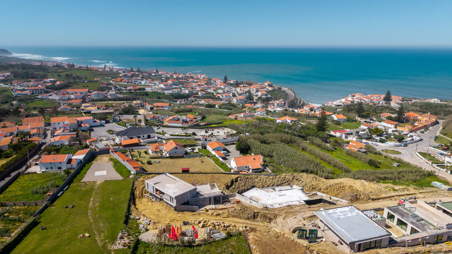 Aerial view of a coastal town with orange-roofed houses, green hills, and blue ocean. New construction sites are visible in the foreground.