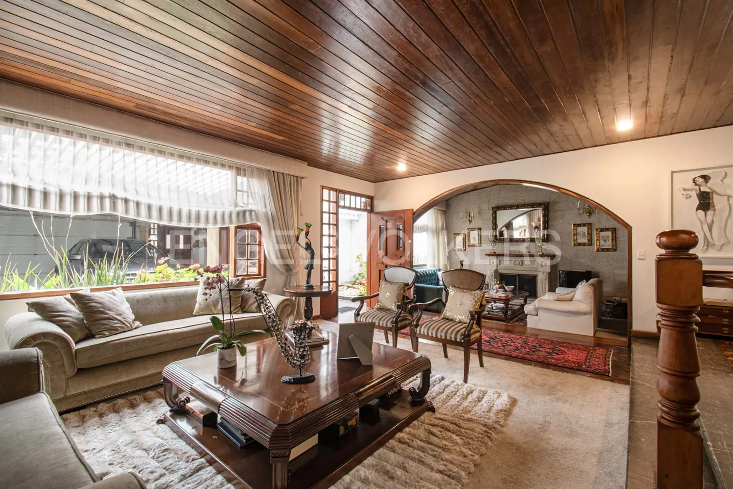 A living room with wood-paneled ceiling, sofas, chairs, and a large window. A doorway leads to an outdoor area.