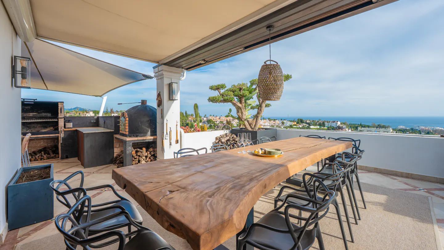 Outdoor dining area with a long wooden table, black chairs, and a view of the ocean. There is a grill and pizza oven in the background.