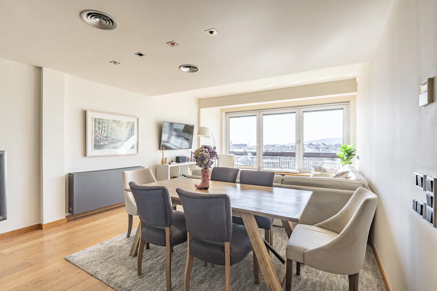 Bright, modern dining area with a wooden table, gray chairs, and beige chairs. A pink boot vase holds flowers. Large window with city view.