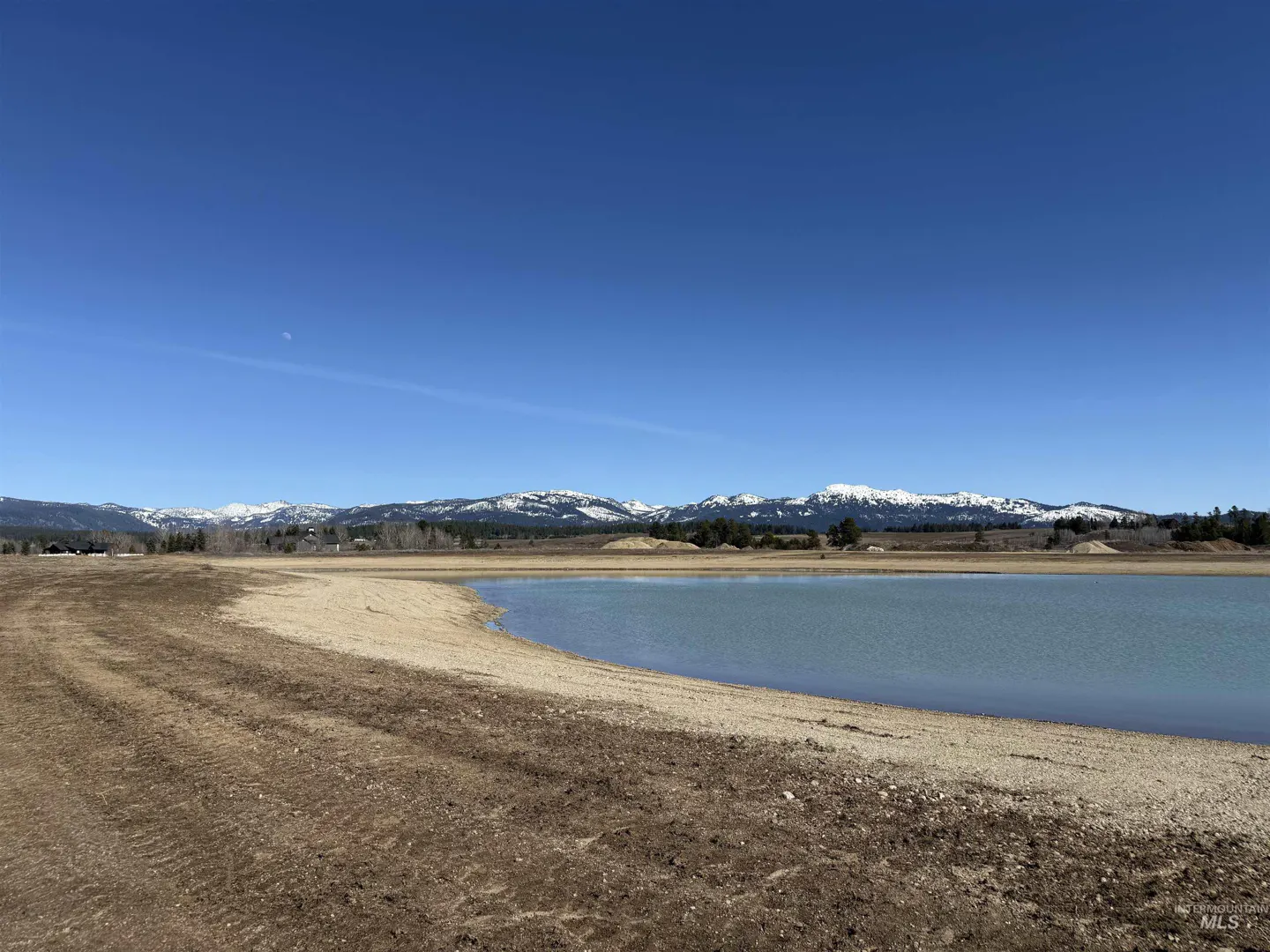 A landscape view of a lake with snow-capped mountains in the background under a clear blue sky.