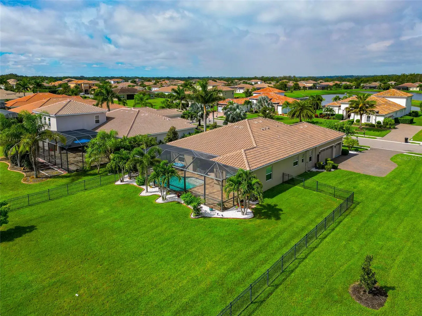 Aerial view of a beige single-family home with a brown tile roof, screened pool, green lawn, and black fence in a residential neighborhood.