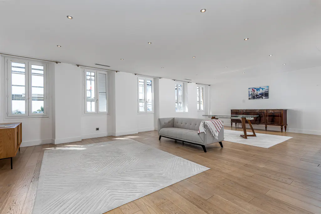 Bright, airy living room with hardwood floors, white walls, and large windows. A gray sofa sits on a patterned rug, with a wooden cabinet and table in the background.