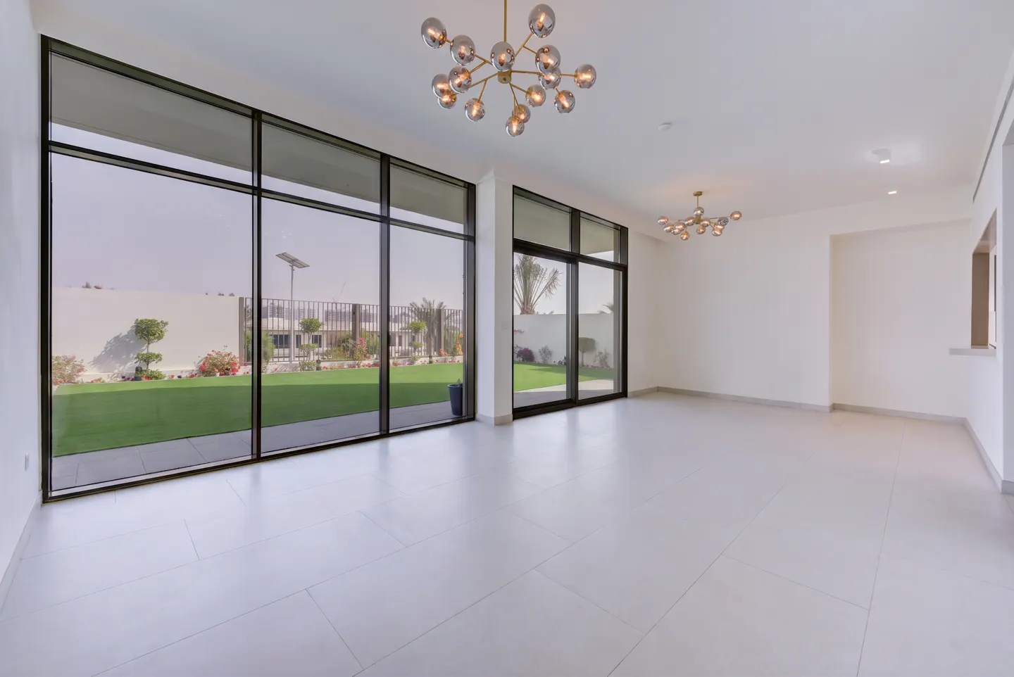 Bright, empty room with white walls, light tile floor, and modern chandeliers. Large windows overlook a green lawn and backyard.