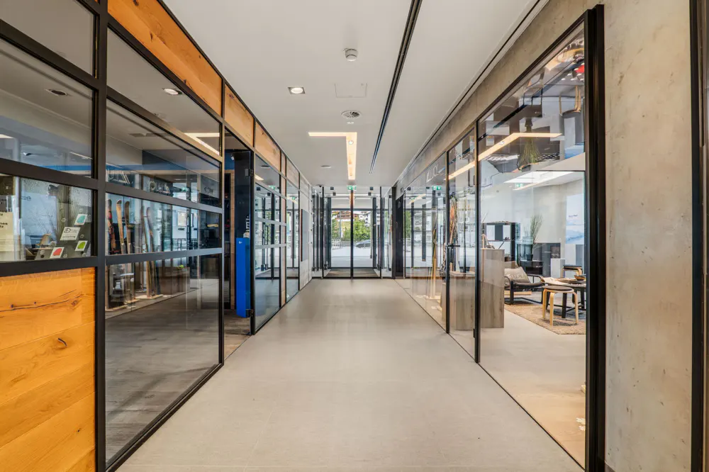 Modern office hallway with light gray floors, concrete walls, and glass-walled rooms featuring wood accents and furniture.