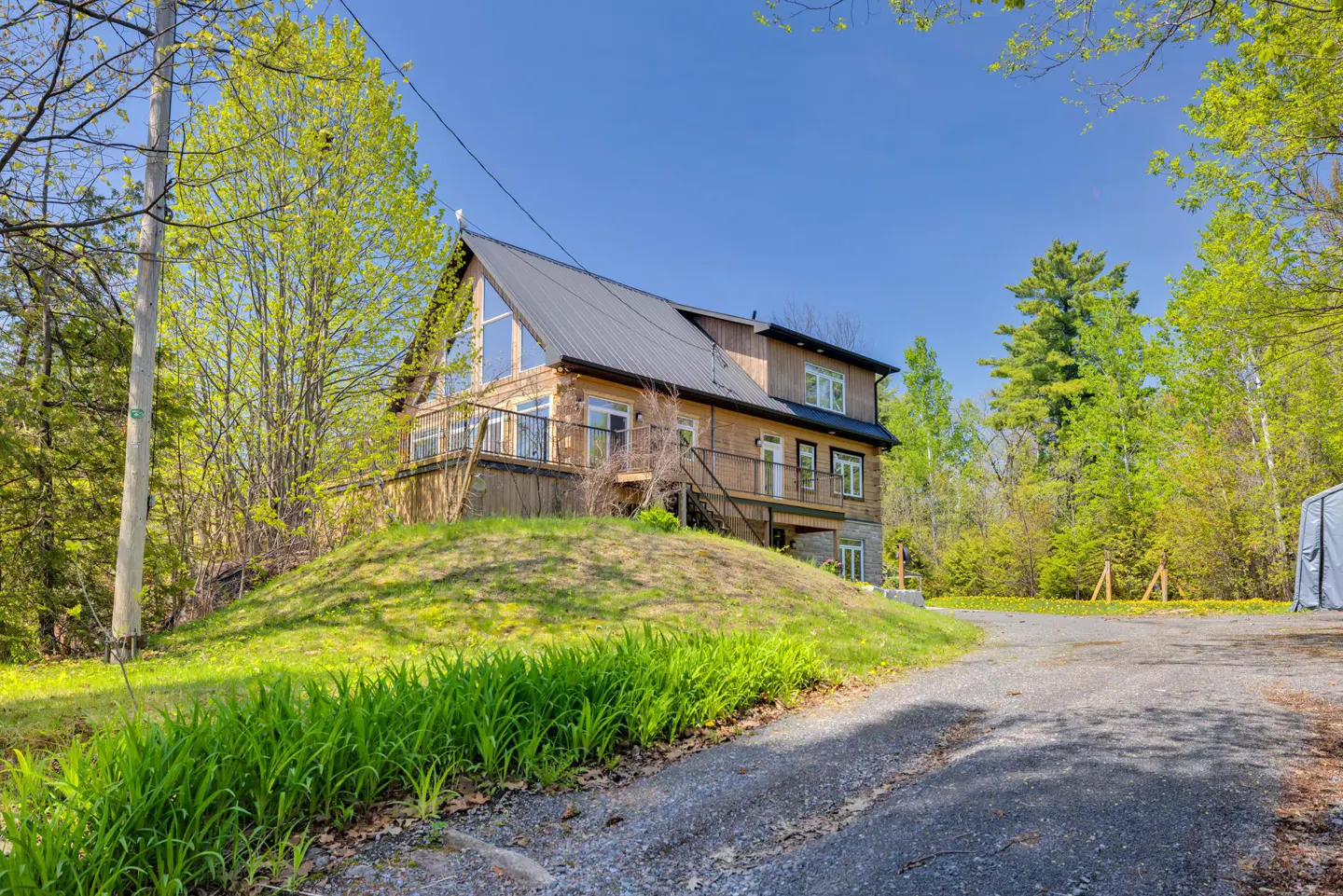 Two-story house with a black roof, wood siding, and a stone foundation on a grassy hill. A gravel driveway leads to the house.