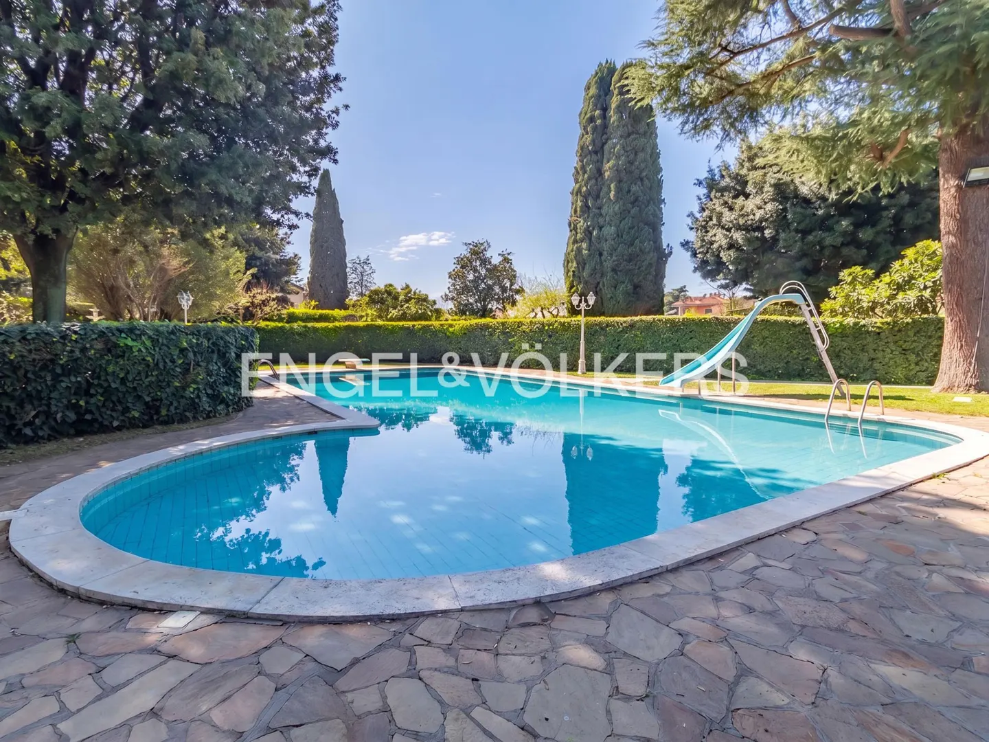A kidney-shaped swimming pool with a blue slide, surrounded by a stone patio, lush green hedges, and tall cypress trees.