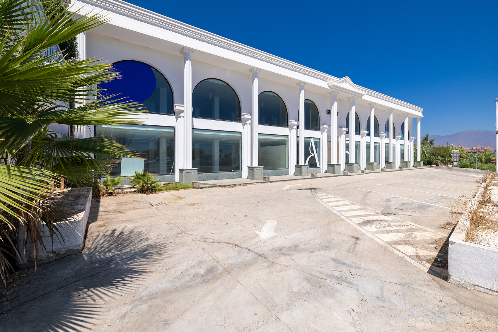 White commercial building with arched windows and columns, featuring a paved parking lot under a clear blue sky. [AI-generated text]