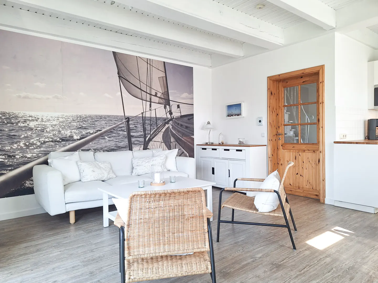 Bright living room with a white sofa, table, and a large sailboat mural. Wicker chairs and a wooden door add warmth to the space.