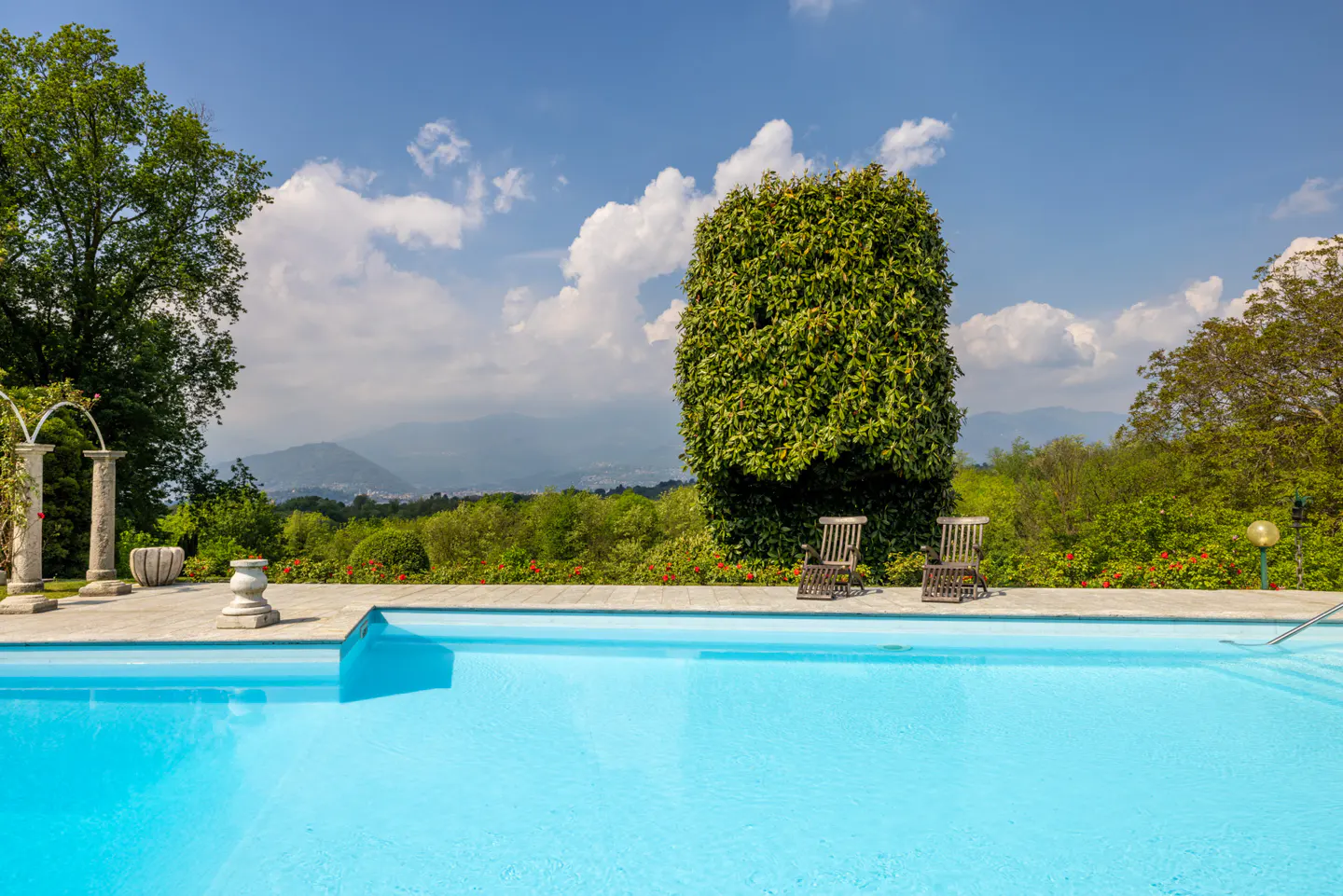 A bright blue swimming pool with two wooden chairs and green trees under a blue sky.