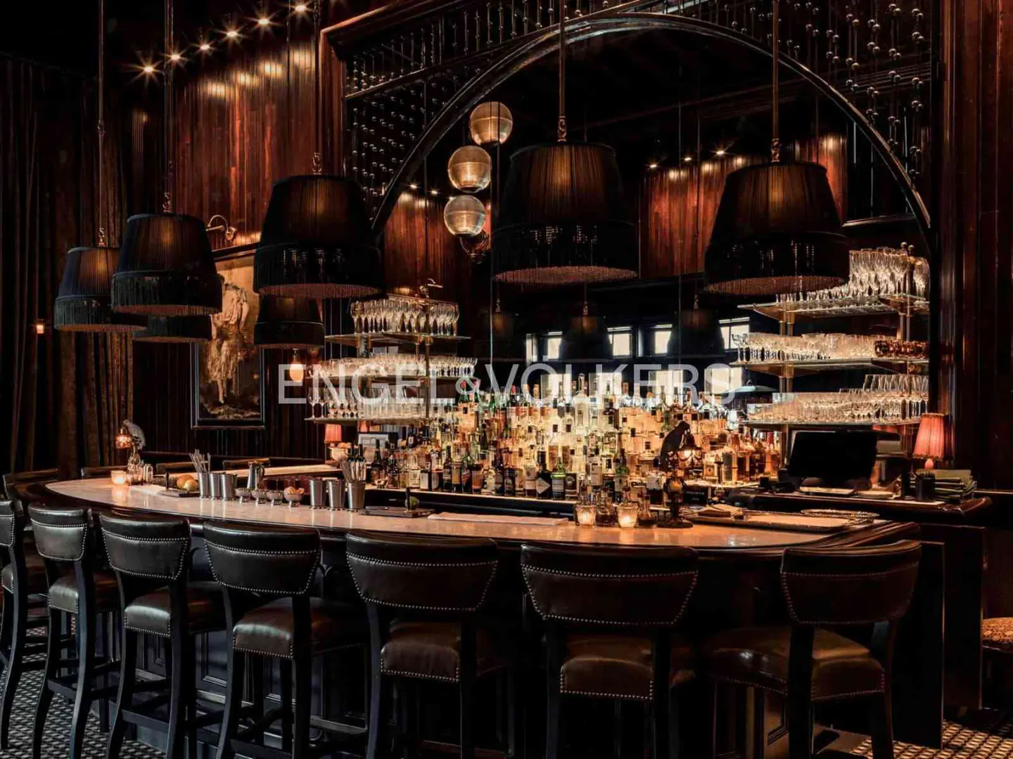 A dimly lit bar with a marble countertop, leather chairs, and black pendant lights. Liquor bottles line the back wall, reflected in a large mirror.