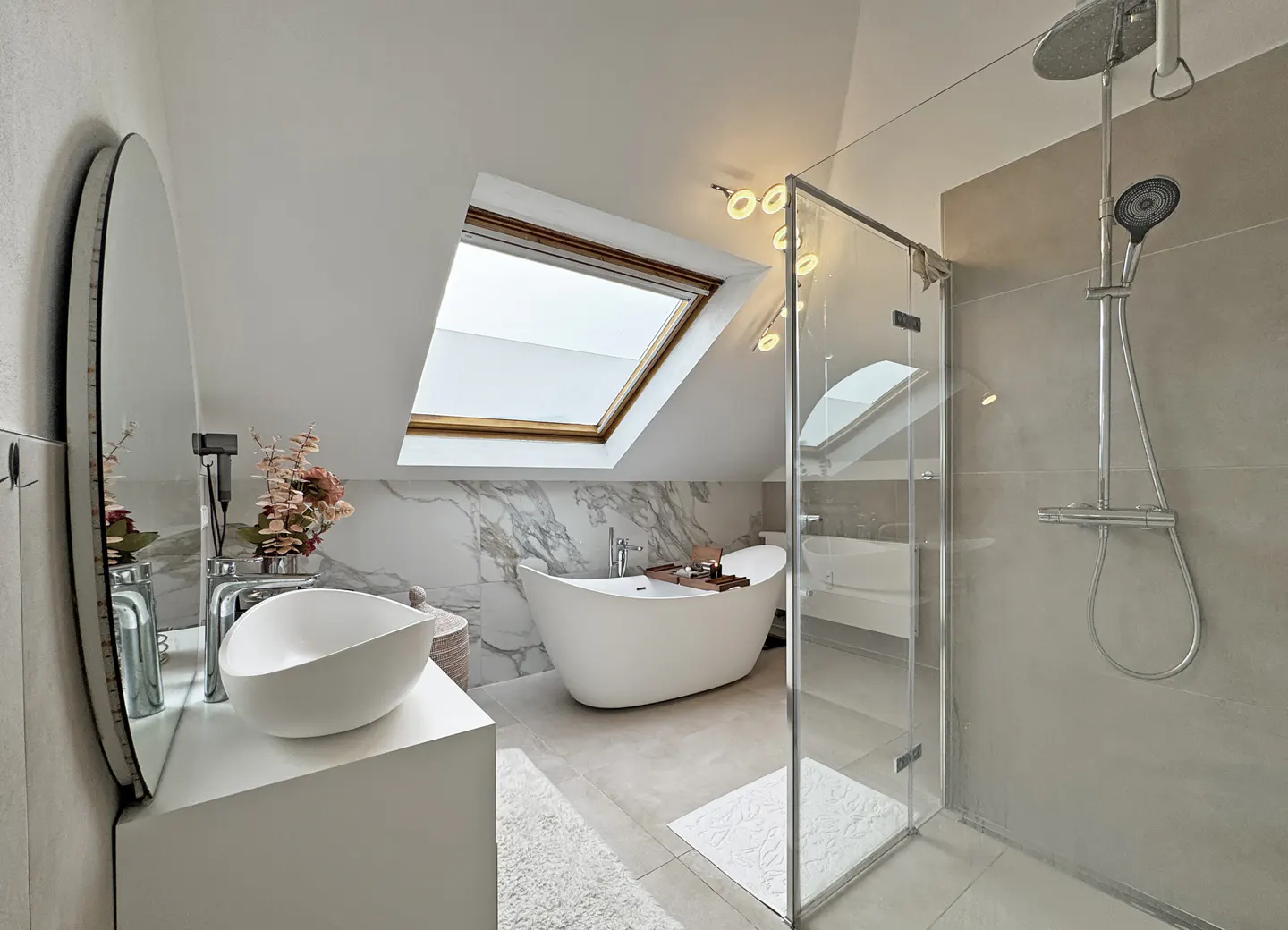 Bright bathroom with white tub under skylight, glass shower, and modern sink with oval mirror.