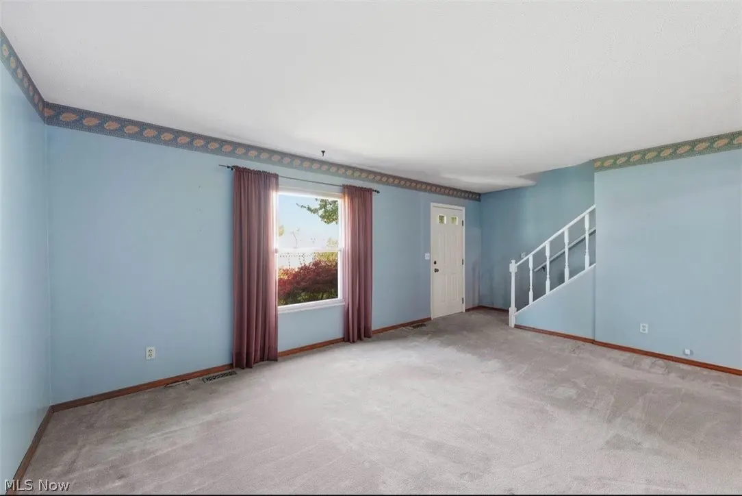 Empty living room with light blue walls, gray carpet, and white trim. A window with maroon curtains and a white front door are visible.