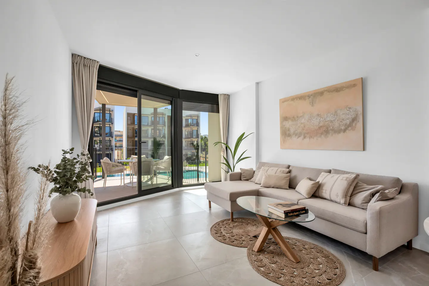 Bright living room with beige sectional sofa, glass coffee table on jute rugs, and a balcony overlooking a pool.