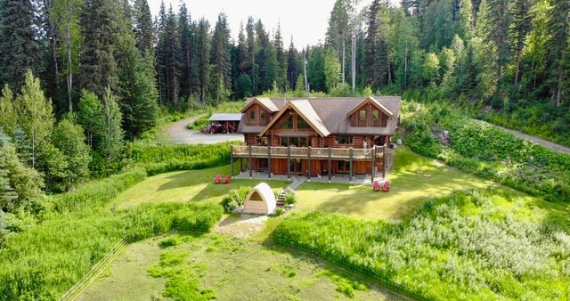 A large brown wooden house with a deck sits in a lush green meadow surrounded by a dense forest of tall pine trees. [AI-generated text]