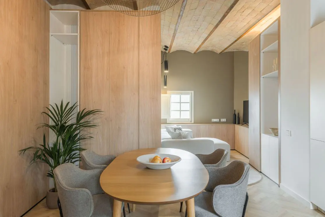 Interior view of a modern apartment with a dining table, chairs, and a glimpse into the living room with a white sofa.