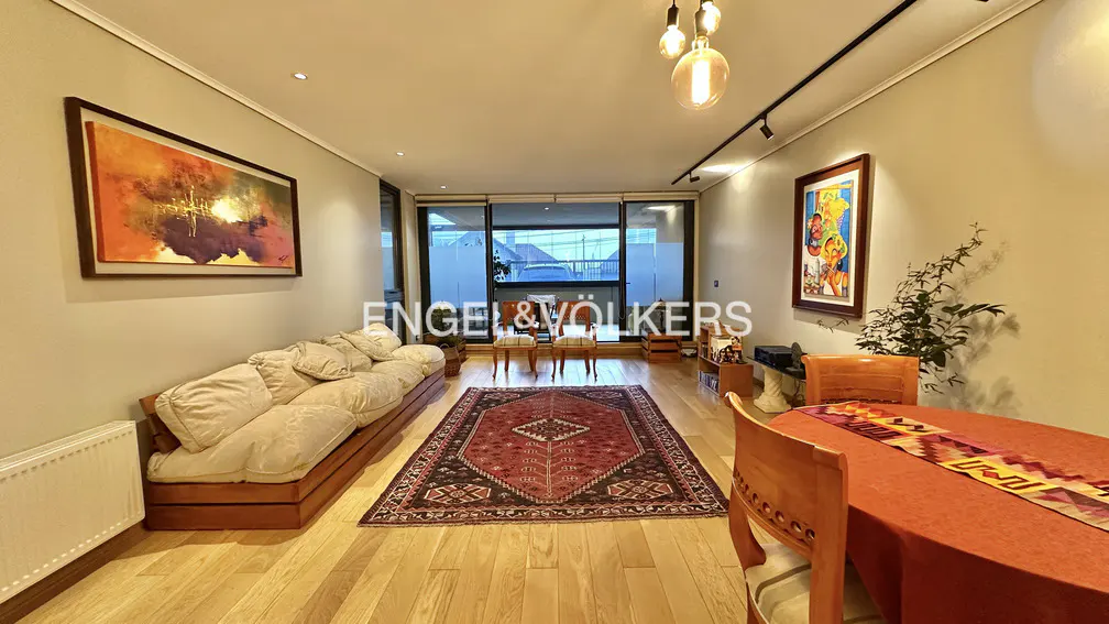 Bright living room with hardwood floors, a beige sofa, and a red patterned rug. A dining table with orange tablecloth is on the right.