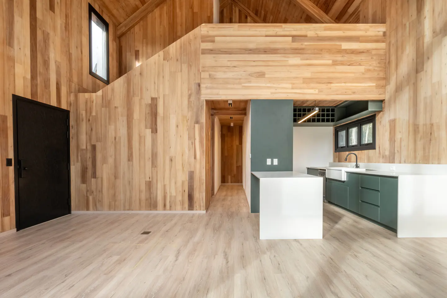 Modern kitchen with light wood walls, a white island, sage green cabinets, and a black door in an open-plan home.