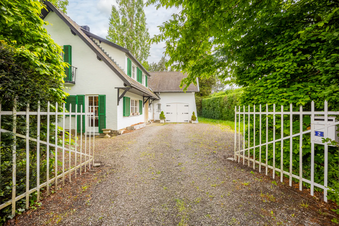 View of a white house with green shutters, seen through an open white metal gate. A gravel driveway leads to a detached garage.