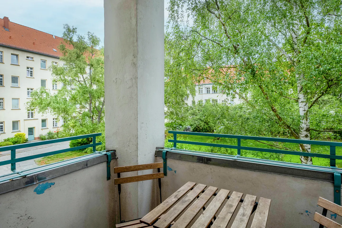 A balcony with a wooden table and chairs, overlooking a green courtyard and a white apartment building with a red roof.
