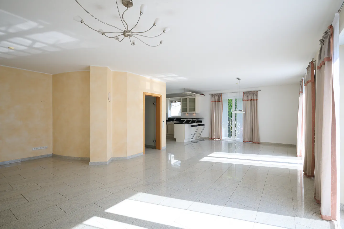 Spacious living room with light yellow walls, white tile floors, a modern chandelier, and an open kitchen in the background.