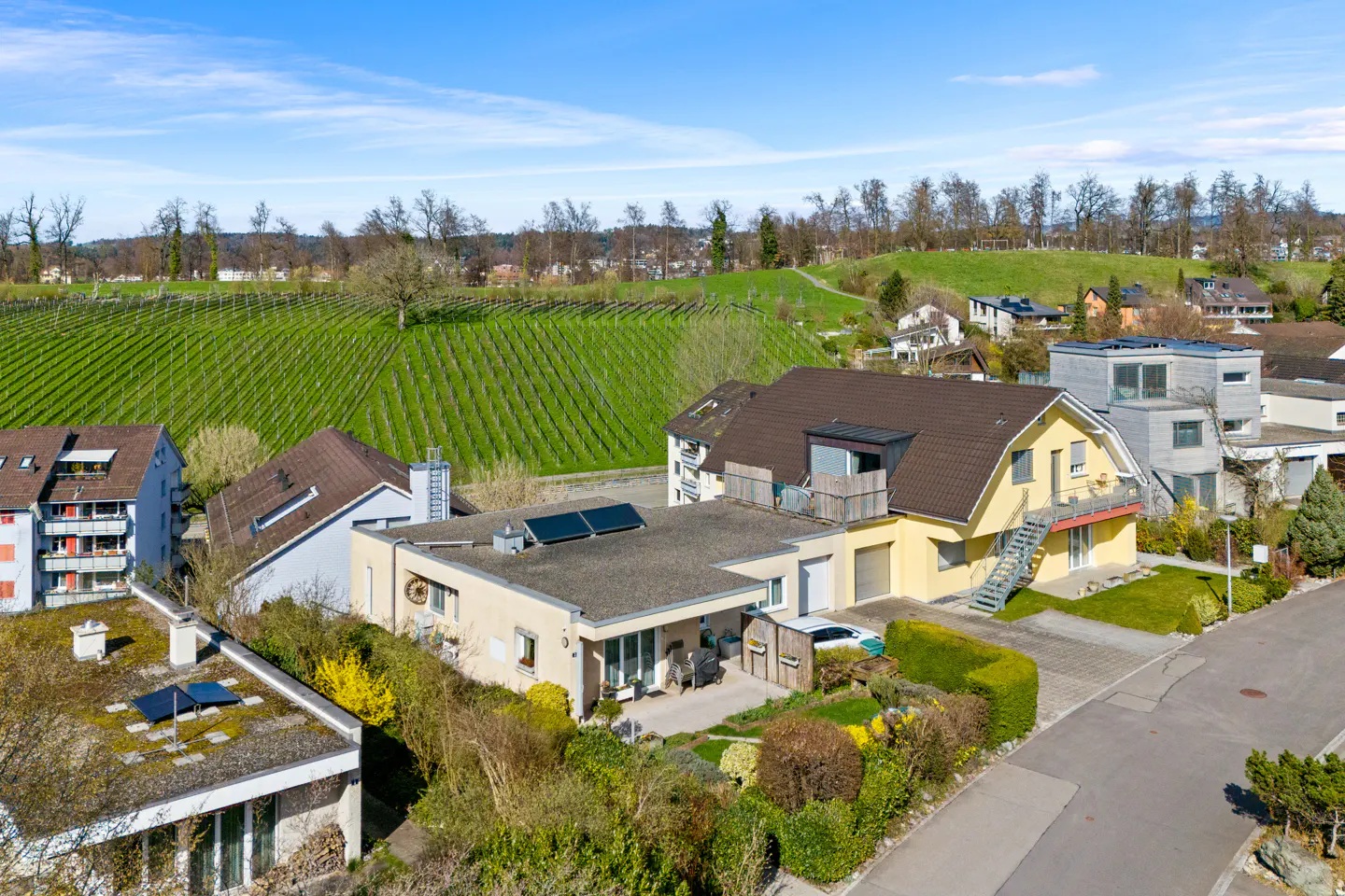 Aerial view of a yellow house with a brown roof, surrounded by greenery and a vineyard in the background under a blue sky.