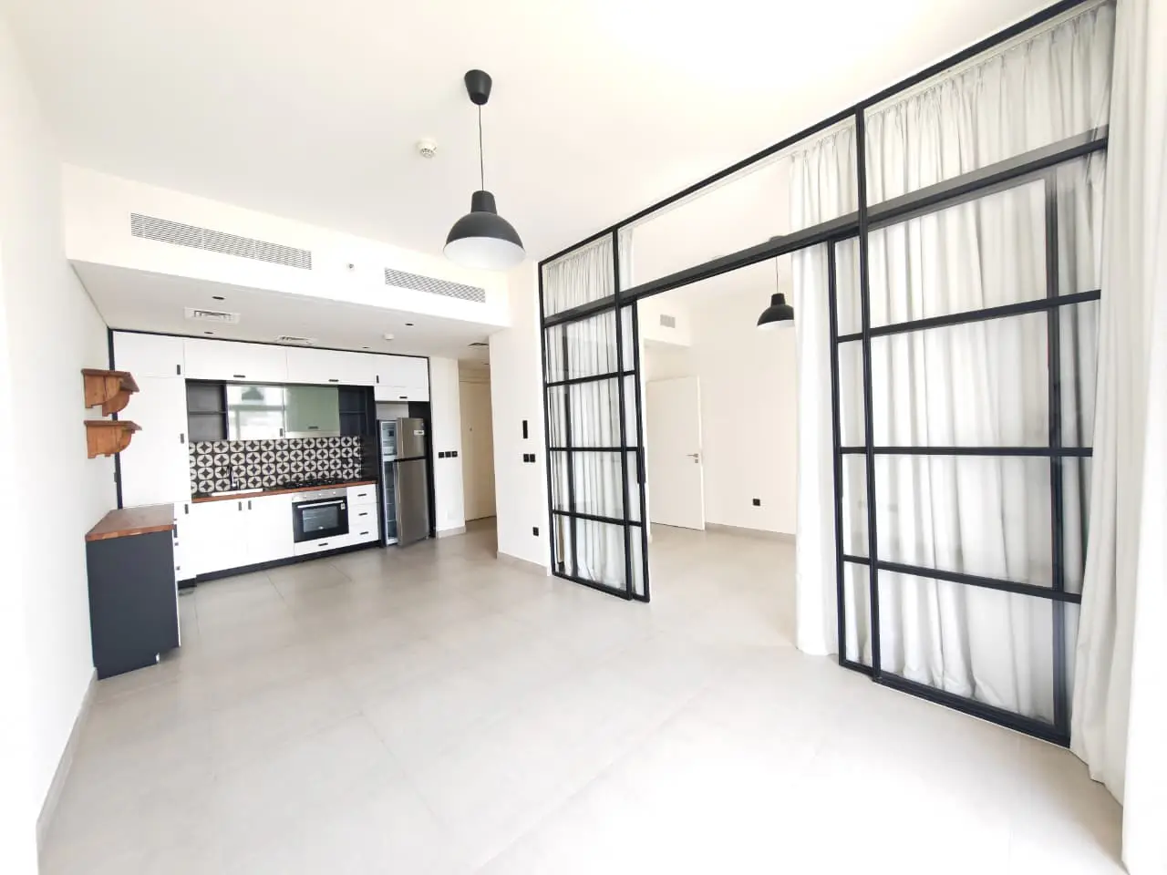 Bright, modern apartment interior with white walls, gray floors, and a black-framed glass partition with white curtains. Kitchen with white cabinets and stainless steel appliances.