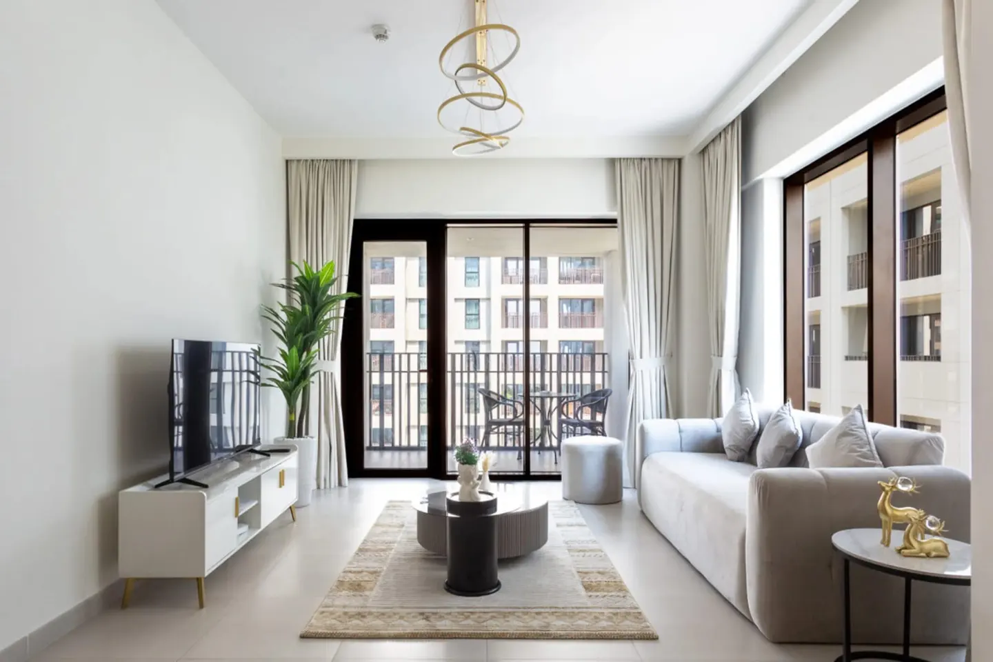 Bright living room with a gray sofa, TV, and balcony. A modern chandelier hangs from the ceiling.