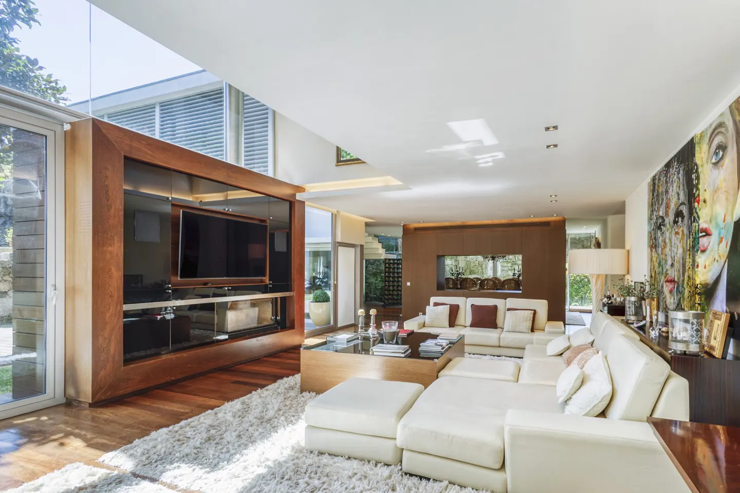Modern living room with a white sectional sofa, glass coffee table, large TV in a wood unit, and colorful wall art.