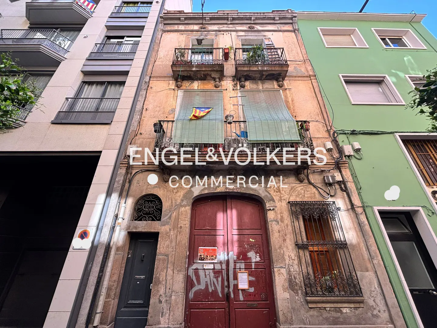 Exterior of a narrow, weathered building with red doors and balconies, flanked by modern buildings. "Engel & Völkers Commercial" overlay.
