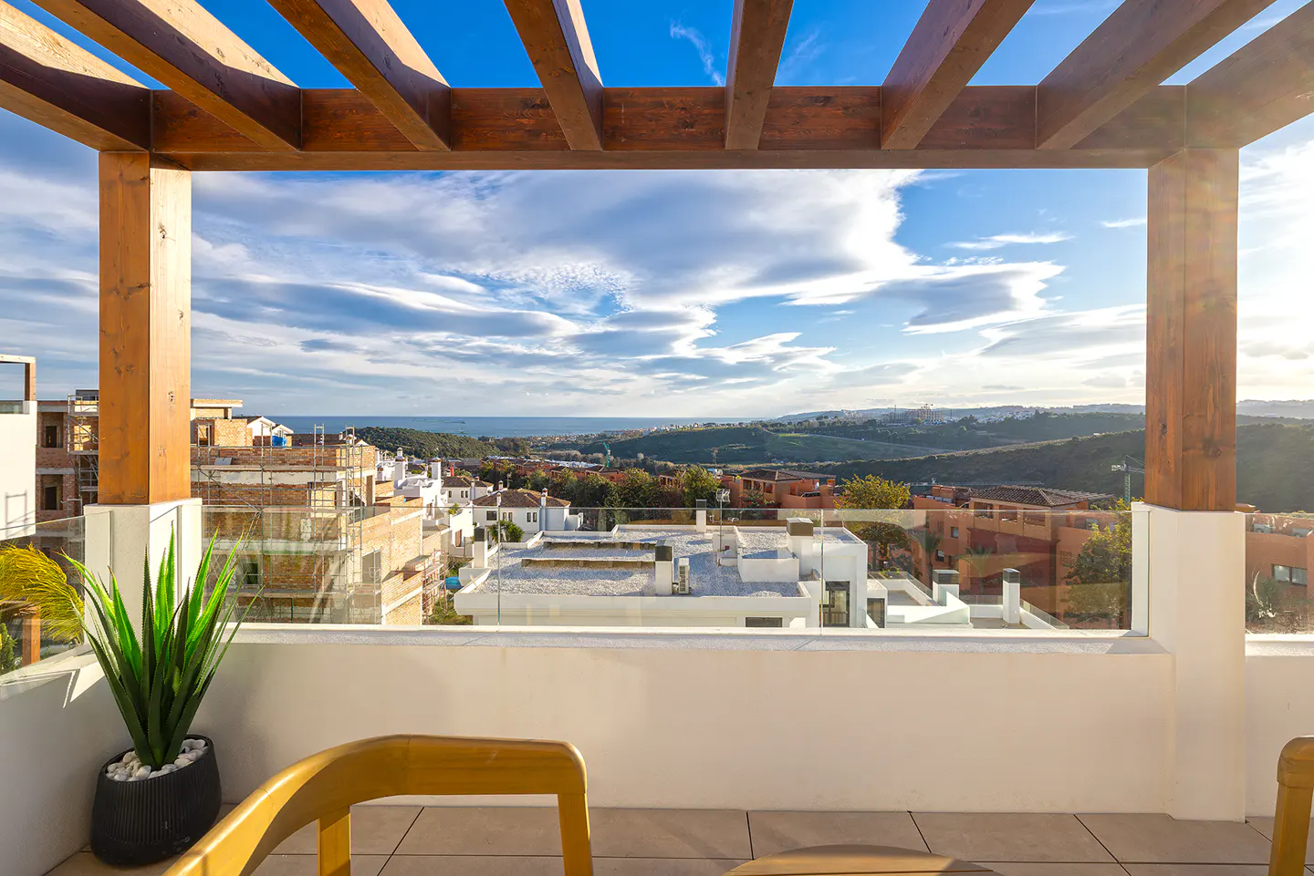 Balcony view with wooden pergola, white railing, and potted plant. Distant cityscape and ocean under a blue, cloudy sky.