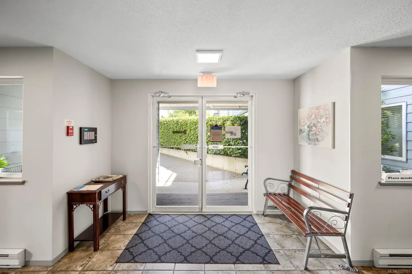 Building lobby with double glass doors leading outside. A bench sits to the right, and a table to the left. A patterned rug lies on the tiled floor.
