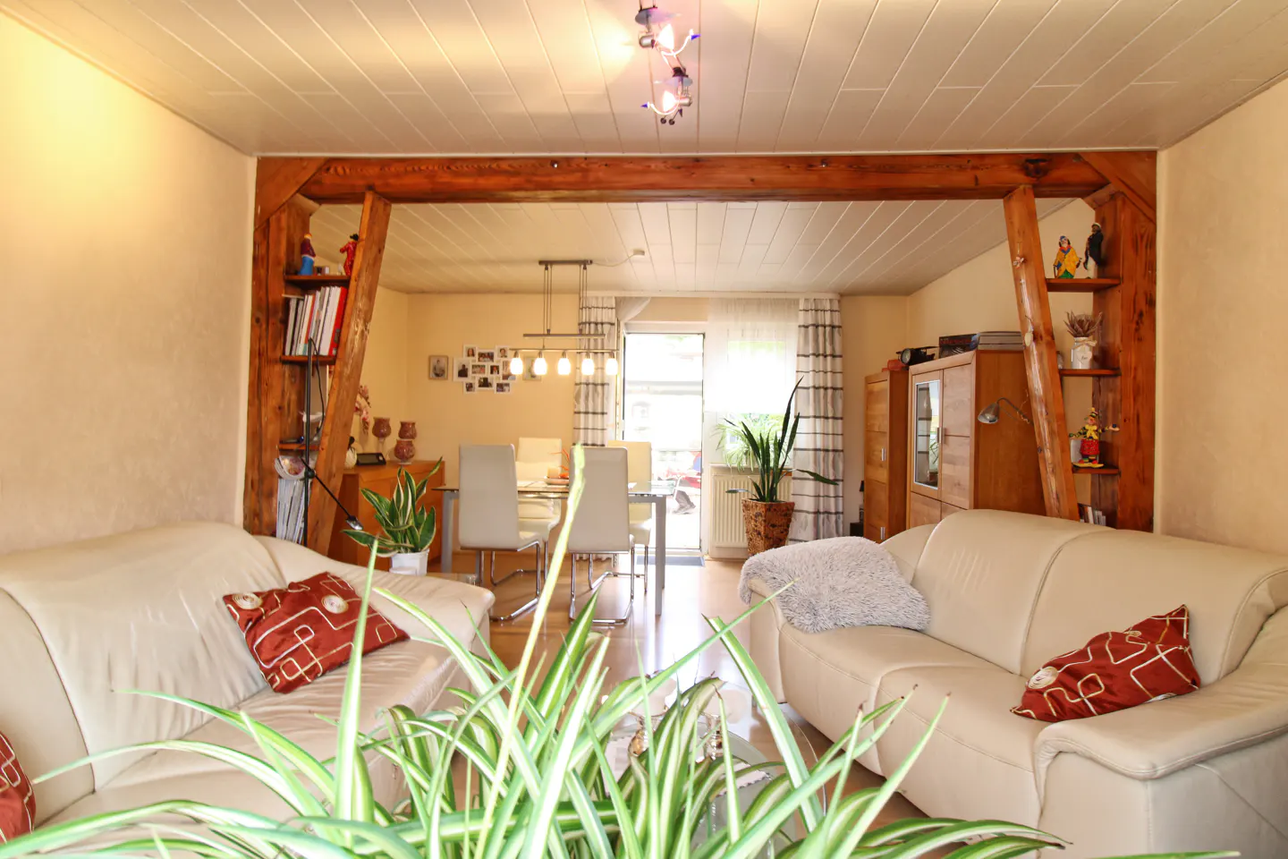 Living room with two cream leather sofas, red patterned pillows, and a dining area visible through a wooden frame.