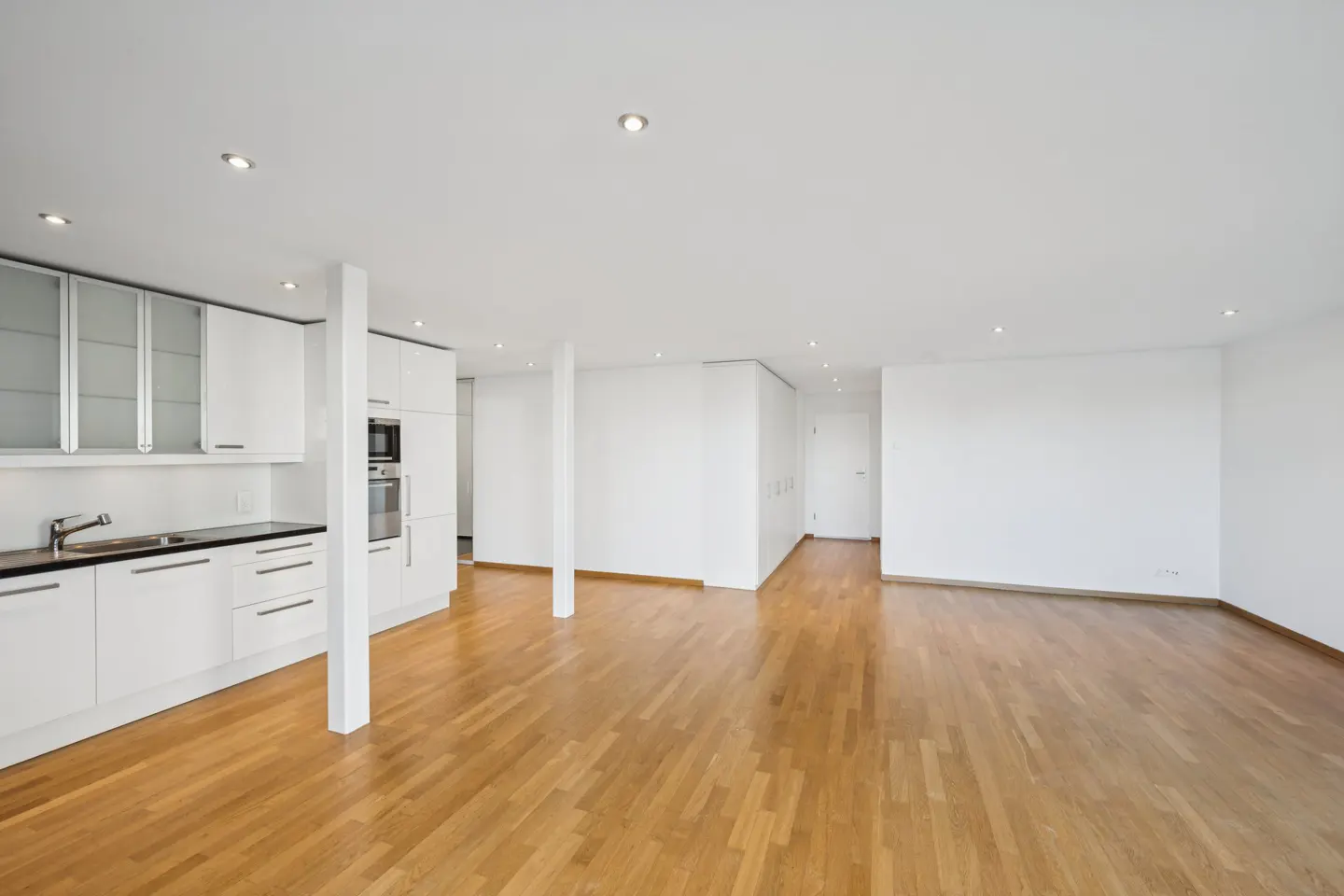 Bright, open-concept apartment with hardwood floors, white walls, and recessed lighting. A modern white kitchen is visible on the left.