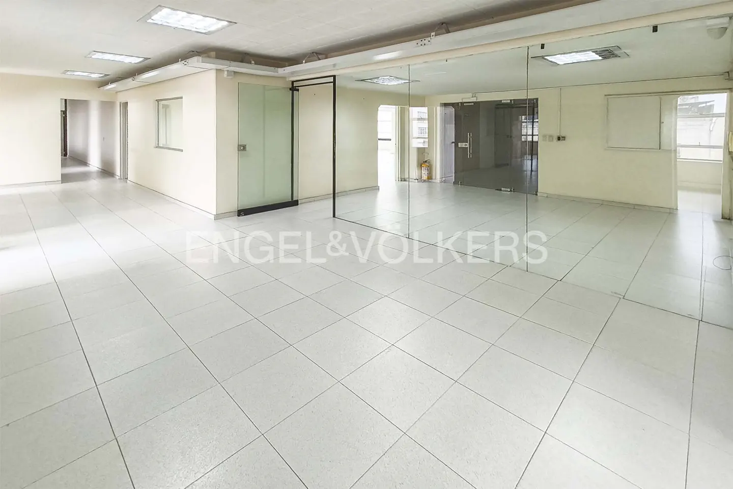 Bright, empty office space with white tile floors and glass-walled rooms. The walls are off-white, and there are fluorescent lights on the ceiling.
