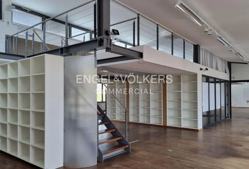 Interior view of a commercial space with a mezzanine, white shelving units, and wooden floors. Stairs lead to the upper level with glass railings. "Engel & Völkers Commercial" logo visible.