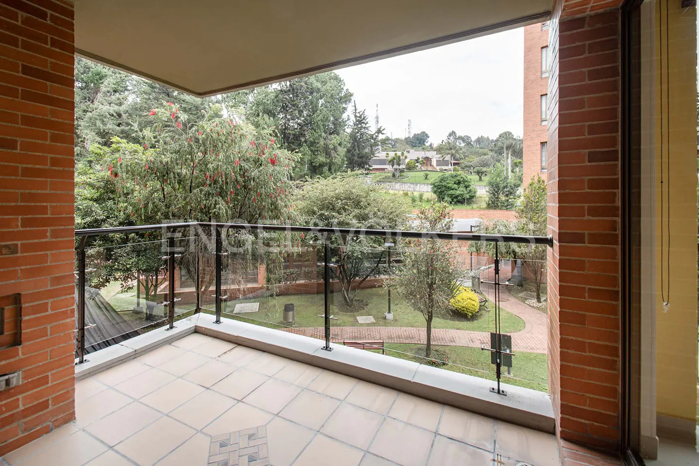 Balcony view with brick walls, tile floor, and glass railing overlooking a green garden with trees and a walking path.