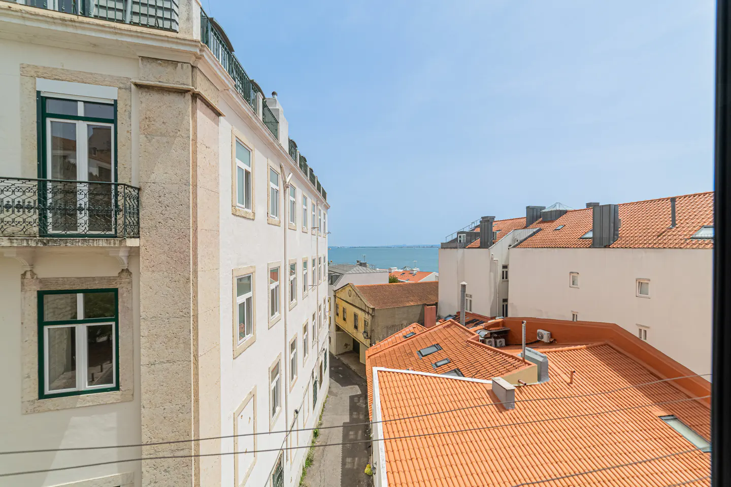 View from a window of white buildings with red tile roofs, a narrow street, and the ocean in the distance under a blue sky.