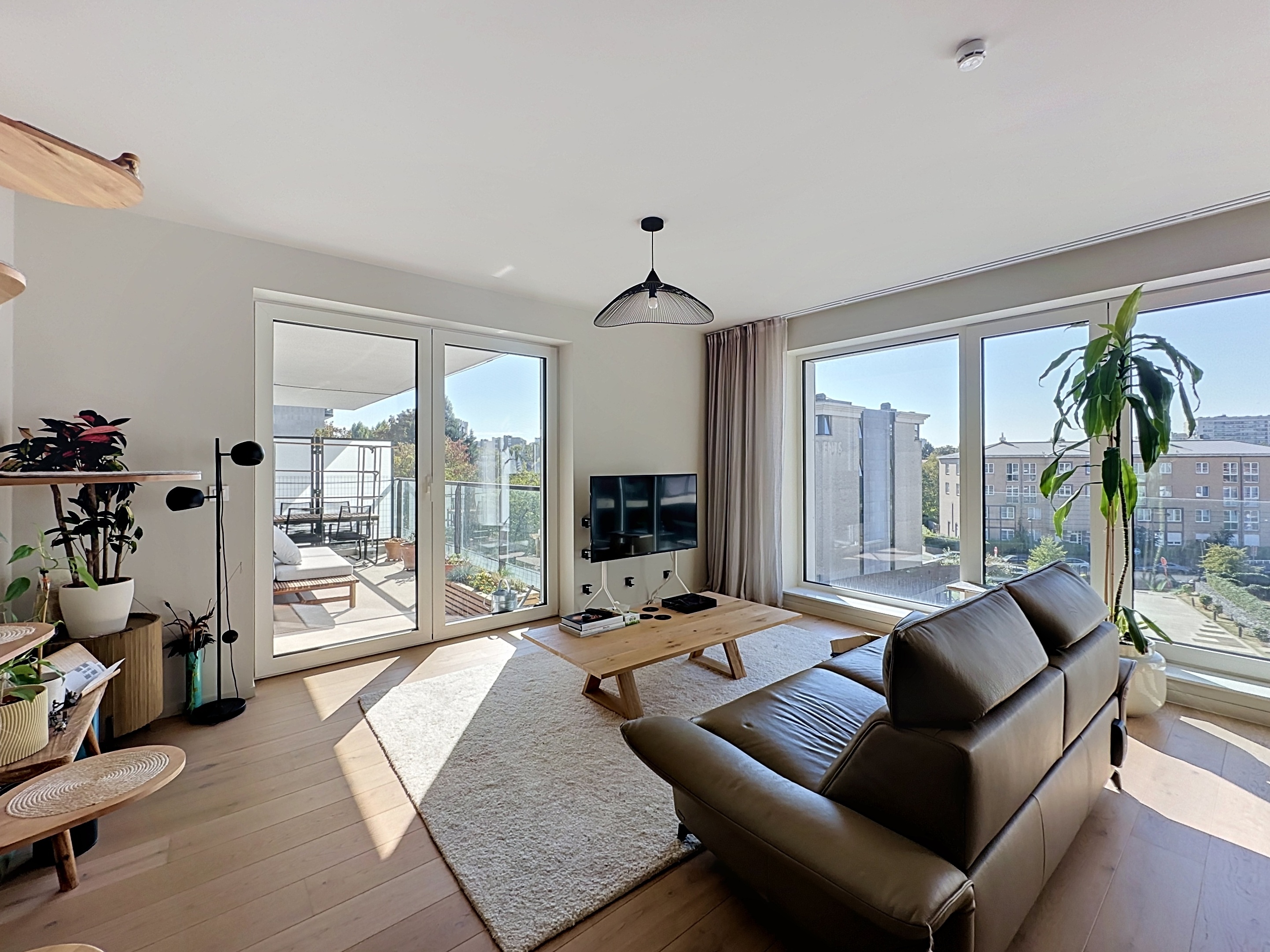 Modern living room with a brown leather sofa, wooden coffee table, large windows, and a balcony with outdoor furniture. [AI-generated text]