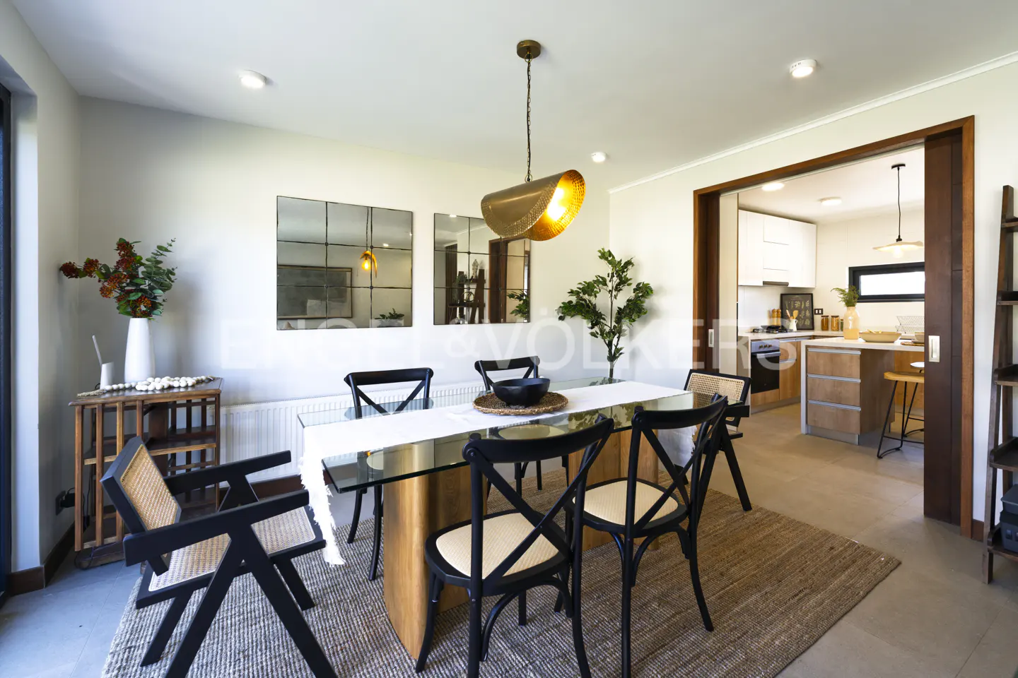 A dining room with a glass table, black chairs, and a gold pendant light. A kitchen is visible through a doorway.