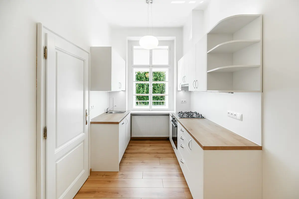 Bright, narrow kitchen with white cabinets, wood countertops, and wood floors. A window shows green foliage outside. A white door is on the left.