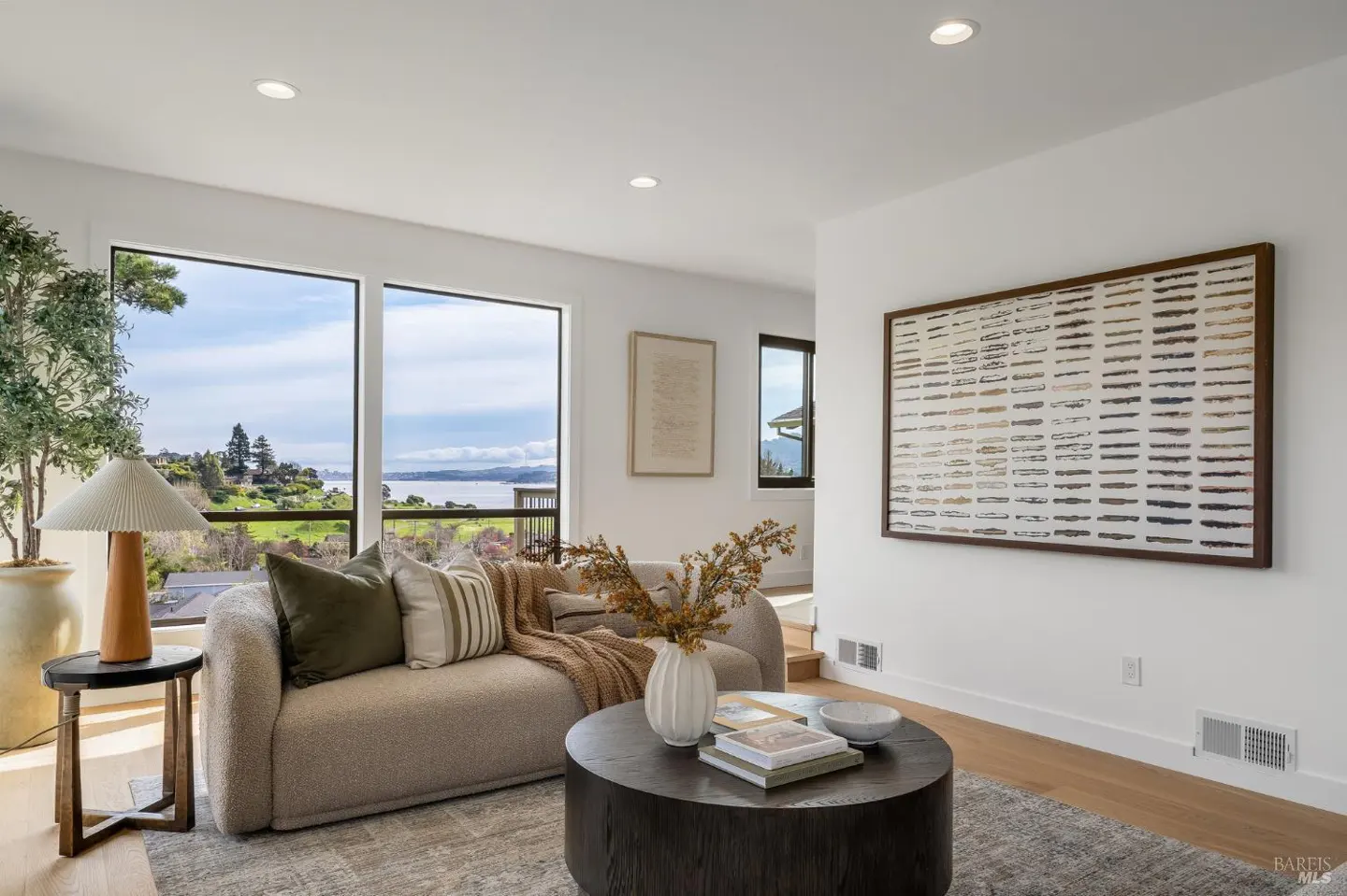 Bright living room with a beige sofa, round coffee table, and large windows overlooking a green landscape and bay. Artwork hangs on the white walls.